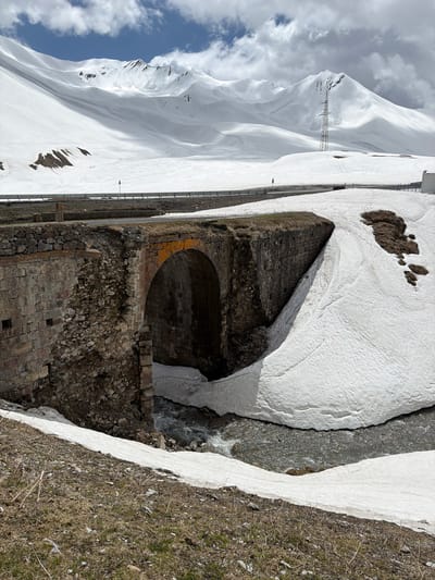 The image shows a dilapidated brick structure partially covered in snow in Ganisi, Georgia. A stream flows next to the structure, with snowdrifts lining its banks. In the background, a snow-covered mountain range dominates the horizon under a cloudy sky. An electrical transmission tower is visible on the mountain slope. The foreground is a mix of brown grass and remaining patches of snow.