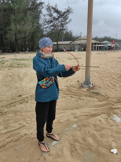 An adult female stands on a sandy beach or coastal area in Thuận An Ward, Vietnam, under an overcast sky. She is positioned facing slightly right, smiling subtly, and holding a reddish-brown, natural-material bow with a string that has a small, light-colored decorative element near its top. Her right hand holds the bow, and her left hand grips a straight wooden stick serving as an arrow, pointing forward. The woman wears a blue and white patterned beanie, a teal long-sleeved jacket, a multicolored fanny pack across her front, black pants, and black thong-style sandals. Her hair is light-colored and styled in two braids on the sides of her head. To her right, a tall, light brown metal pole with a grey bolted base extends vertically from the sand. The ground is composed of light brown sand exhibiting footprints and scattered small pieces of white debris. In the background, a line of tall, slender trees with green foliage provides a backdrop. Beyond the trees and to the right, multiple low-rise buildings are visible, including simple structures and larger commercial buildings, one featuring a multi-tiered roof and signage that partially displays "SAIGON." The sky is uniformly grey and cloudy.