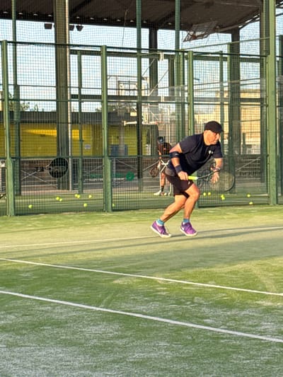 A man wearing a black t-shirt, dark shorts, a black cap, purple athletic shoes, and a black arm brace on his left arm is playing padel on an outdoor court. He is in mid-action, bending his knees and holding a padel racket in his right hand, poised to strike a yellow ball. Several yellow padel balls are scattered on the green court surface behind him. The court is enclosed by green metal fencing with netting. In the background, another person is visible, also holding a racket. The court is situated within a larger structure with a metal roof and vertical supports, suggesting it's part of a sports complex in Vidreres, Spain. The lighting indicates it is daytime, with sunlight casting shadows on the court.