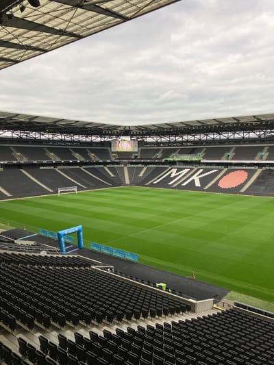 The image shows a partial view of Stadium MK in Milton Keynes, United Kingdom. The stadium is largely empty, save for a few individuals near the field. The green field is striped with slightly darker lines. Blue arches labeled "MK Finish 5K" are placed near the edge of the field. A goal post is visible near the far end of the field. Black seats occupy most of the visible stands, with some white and red decorative elements on one side. The sky is overcast. Structural elements of the stadium roof are visible on both sides of the frame.