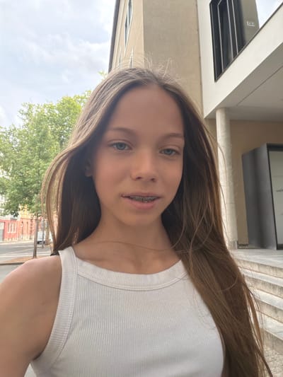 The image shows a young person, likely a girl, with long brown hair and blue eyes, wearing a white tank top and braces. They are standing outdoors in what appears to be an urban environment. Behind the person, there is a building with a modern architectural design, characterized by clean lines and large windows. To the left, there are trees and buildings that suggest a street or a sidewalk with some traffic. The sky is cloudy, indicating an overcast day. The scene is likely a public space in the city of Würzburg, Germany, given the prompt's instruction to include this location. The overall impression is a casual, candid portrait taken outside.