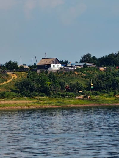 The image shows a rural landscape along a body of water, likely a river, in Erkalovo, Russia. The foreground is dominated by the rippling surface of the water. The midground features a grassy bank with several structures on a sloping hill above it.
The most prominent structure is a dark wooden house with a white section and a peaked roof. Adjacent to it is a greenhouse with a rounded translucent roof. Further down the slope, a small, ornate golden-domed building, possibly a chapel, is visible amidst the greenery. There are also a few scattered animals, appearing to be cows, grazing in the field. A dirt path winds its way up the hill.
In the background, trees and other smaller buildings create a rustic, village-like setting. The sky is a pale blue with a few wispy clouds. The overall scene depicts a tranquil, rural environment.