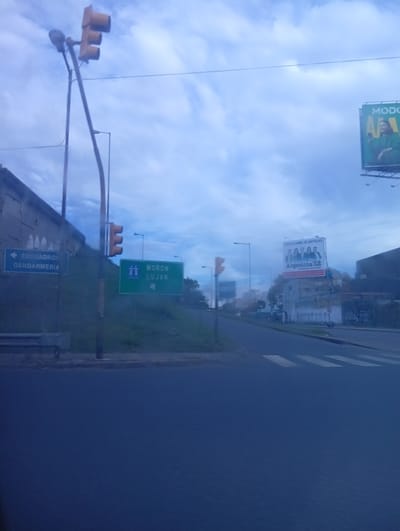 The image shows a street scene in Ciudadela, Argentina. A traffic light is visible, along with street signs indicating directions to "ESCUADRON GENDARMERIA" and "MORON LUJAN". There are billboards on the right side of the street, one advertising "MODO" and another for "Argentina 12". The sky is cloudy, and the street is wet. The photograph appears to have been taken from inside a vehicle. A crosswalk is visible.