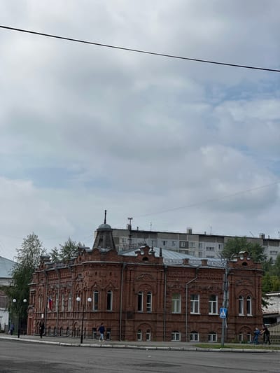 The image shows a large, ornate, two-story brick building with a complex roofline and multiple decorative elements. The building is made of red brick and has numerous windows, some arched. A Russian flag is visible on the front of the building. Several people are walking on the sidewalk in front of the building, and some are near the street. In the background, there is a multi-story apartment building and trees. The sky is overcast. The scene appears to be located in the city of Nogornyi, Russia.