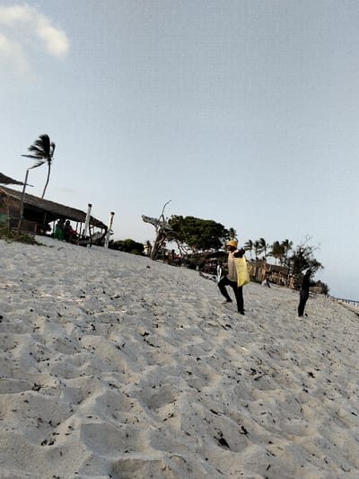 The scene depicts a sandy beach or dune environment in Gombato Bongwe ward, Kenya, viewed from a low angle looking uphill. The foreground and mid-ground are dominated by light-colored sand, showing numerous footprints and scattered dark organic matter.

In the mid-ground, a person wearing a yellow hat, a light-colored long-sleeved shirt, dark pants, and dark footwear is walking uphill from the right side of the frame towards the left. A light yellow or beige bag is carried on their back. To their right, further uphill, another individual is partially visible, dressed in dark clothing.

In the background on the left, a structure with a distinct thatched roof is visible, supported by wooden poles. Underneath this open-air structure, several individuals are seated. Adjacent to the structure, a tall palm tree with fronds bending left, indicative of wind, stands against the sky. Further right in the background, a dense cluster of green foliage and additional palm trees are present. A light-colored wooden or driftwood-like structure with illegible text (partially visible as "WAT") is positioned near the foliage.

The sky is a light, clear blue, with a single small white cloud visible in the upper left quadrant. The overall lighting suggests either early morning or late afternoon.