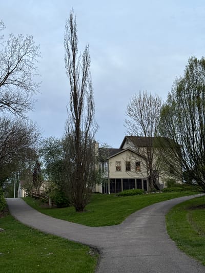 The image depicts a residential scene in Grove City, Ohio, USA. A two-story house with a beige exterior and a screened-in porch is visible. Bare trees surround the house, suggesting late autumn or early spring. A paved pathway curves through the green grass next to the house. The sky is overcast, and the overall lighting is dim. The scene appears quiet and residential.