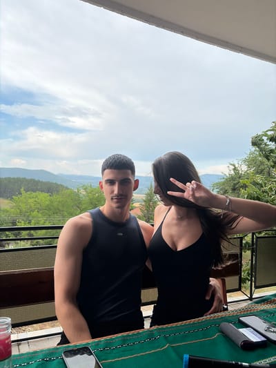 The media file shows two young adults, a woman and a man, on a balcony overlooking a lush, mountainous landscape. The woman, with long dark hair, is leaning over and making a peace sign with her right hand, her face obscured. She is wearing a black tank top and a silver bracelet. The man, in the foreground, is looking directly at the camera with a neutral expression. He has short, dark hair and a mustache, and is wearing a dark tank top. Behind them, the landscape is green with trees and rolling hills, suggesting a rural or mountainous location. The sky is partly cloudy, indicating daytime. On the table to the right, a green cloth with an embroidered pattern is visible, along with a smartphone displaying the text "TEREA". The overall scene suggests a casual outing or vacation. The presence of "TEREA" on the phone, combined with the location context of Vladislavtsi, Bulgaria, may indicate a connection to a specific product or service available in that region.