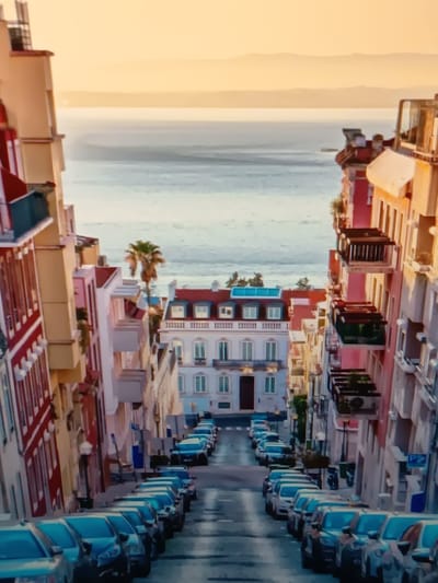 The image depicts a steep, narrow street lined with colorful buildings, descending towards a body of water and a hazy horizon. The architecture suggests a European city, possibly with a Mediterranean influence, characterized by balconies, intricate window frames, and a mixture of stucco and tiled facades in shades of yellow, pink, and red. The street is paved and slopes downwards, with numerous cars parked along both sides.
In the foreground, several blue and silver cars are visible, parked bumper-to-bumper, indicating a dense urban environment. Further down the street, more cars continue the line, their shapes blurred by the distance and resolution. A single palm tree stands out amidst the buildings on the left side of the street, adding an exotic touch.
In the middle ground, a distinctive white building with a red-tiled roof and prominent windows is situated at the end of the street, framing the view of the water. This building features a prominent central section with a series of windows and a balcony, and a smaller section to its right. Above the water, a pale, hazy sky stretches to the horizon, suggesting either dawn or dusk. The water itself appears calm, with subtle ripples.
The overall impression is one of a picturesque, possibly historic, urban landscape. There are no people visible in the image. The scene is static, with no active events occurring. The atmosphere is serene, with the soft light bathing the scene.
This image is not of Serhiivka, Ukraine.