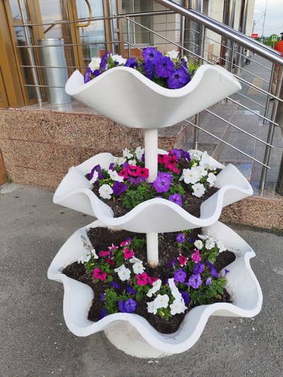 A multi-tiered white planter filled with colorful petunias stands on a paved surface in front of a building. The planter has three wavy levels, each overflowing with flowers in shades of purple, pink, and white. The building features large glass doors and a granite-tiled facade. A metal handrail runs alongside a set of stairs leading up to the building entrance. In the background, a person in an orange shirt and light-colored pants walks away from the camera on a sidewalk. A street sign with Cyrillic lettering is visible in the distance. The scene appears to be set outdoors during daylight hours, likely in a public area. There is no clear indication of the weather. The overall impression is one of urban landscaping and street life.