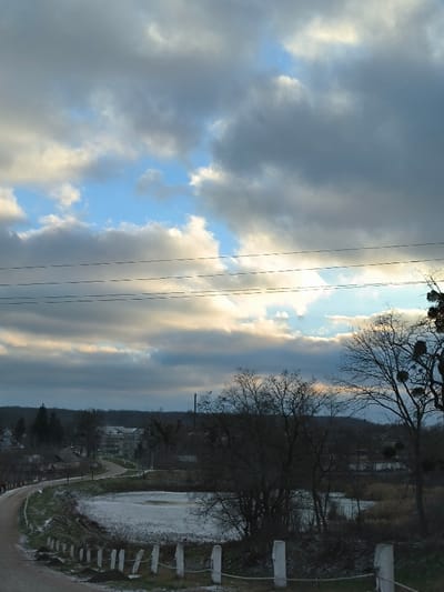 An elevated view captures a wintry landscape in Khrinivka, Ukraine, under a dramatic sky. The foreground features a dirt or gravel road, lightly covered in patchy snow or frost, curving leftwards and descending out of frame. Along the right edge of this road, a low fence composed of regularly spaced white posts connected by a horizontal rope or wire is visible, also dusted with snow. To the right of the road and fence, a partially frozen pond, its surface a dull white, is surrounded by bare deciduous trees and shrubs, some with traces of snow.

In the midground, the road continues to curve, leading towards a cluster of buildings interspersed with trees. Several multi-story structures, possibly residential or institutional, are visible amongst the leafless trees. A tall, slender industrial-style chimney or vent stack rises on the left side of the midground. The background consists of a rolling, tree-covered landscape, with additional scattered buildings.

The sky is heavily overcast with prominent grey and white cumulus clouds. Patches of brighter blue sky are visible through breaks in the cloud cover, particularly towards the upper center and right. Sunlight, possibly from a low-lying sun, illuminates the undersides and edges of some clouds, creating highlights. Multiple utility wires stretch horizontally across the upper portion of the frame. No individuals or active subjects are present in the scene.