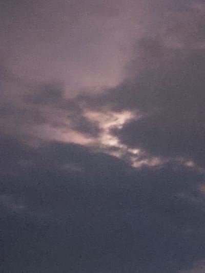 The image shows a cloudy sky with patches of light breaking through. The clouds are a mix of dark gray and lighter shades, with some areas appearing pinkish or orange, suggesting either sunrise or sunset. The overall atmosphere is muted and somewhat hazy, with a grainy texture throughout the photograph. No other elements like buildings, land, or people are visible in this close-up shot of the sky. The context of Kyzylorda, Kazakhstan is not visually discernible from the image itself, as it only captures the sky.