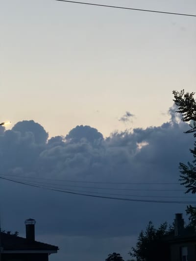 This is a vertical, medium-shot, natural light, outdoor photograph of a sky with some clouds and silhouettes of trees and rooftops. The image is taken during dusk or dawn, as indicated by the soft, pale yellow to light gray gradient of the sky. A thick, dark gray cloud formation stretches horizontally across the middle of the frame, with lighter, fluffier clouds above it. To the left, a bright, yellow light source, possibly the sun peeking through the clouds or a streetlamp, illuminates the edges of some clouds. Several dark, thin electrical wires are strung horizontally across the upper and middle sections of the image. In the lower portion of the frame, the dark silhouettes of a pitched rooftop with a chimney are visible on the left, and on the right, trees and another chimney can be seen against the darkening sky. There are no people present in the image. The scene appears to be an urban or suburban landscape at twilight. The photograph does not contain any specific identifying features to determine its location within the city of Arcahueja, Spain.