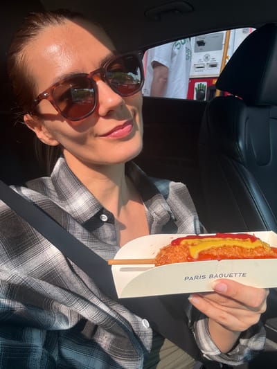 A young woman with light brown hair, wearing sunglasses and a gray and white plaid shirt, sits in the driver's seat of a car. She holds a Korean corn dog topped with ketchup and mustard in a white cardboard container labeled "PARIS BAGUETTE" in her right hand. The background shows a window with a glimpse of a gas station. The scene appears to be in Seorak-myeon, South Korea.