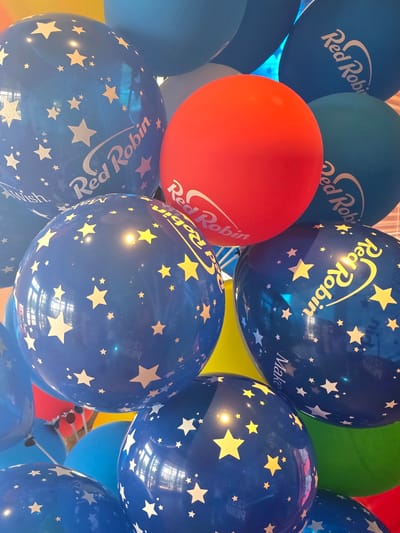 A close-up, vertical, eye-level shot captures a vibrant cluster of Red Robin-branded balloons, predominantly blue and red, with some other colors like green and yellow partially visible. The balloons are decorated with stars, some in white and some in yellow. The Red Robin logo, featuring stylized text and a bird silhouette, is imprinted on several of the blue balloons. The lighting suggests an indoor setting, possibly a party or promotional event, with reflections of the surrounding environment visible on the glossy balloon surfaces. The image does not provide any specific landmarks or contextual clues that would indicate its location in Cumming, Georgia, United States of America. No people are visible in the frame.