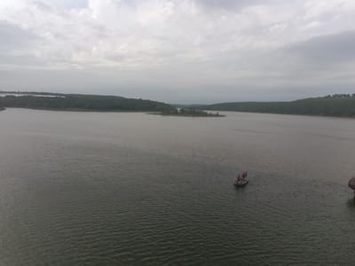 The image shows a body of water, possibly a lake or reservoir, under an overcast sky. Land covered with trees surrounds the water. In the water, there is a boat carrying multiple individuals. The location is likely in a rural or natural area, and I cannot verify if the image is located in the city of Park Hill, United States.
