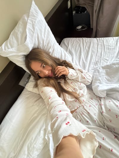 A young woman lies on a bed in what appears to be an interior space, possibly a hotel room or residence. She is wearing a white pajama set patterned with red cherries. The bed is made with white sheets and pillows, and the room appears to be dark. A dark object resembling a suitcase or storage container is visible near the bed. No immediate evidence suggests a specific location in Sosnovy, Russia.