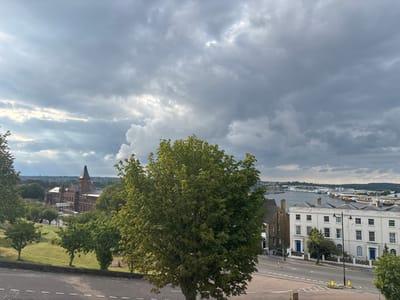 The image depicts an elevated view of a scene in Chatham, United Kingdom, specifically looking towards the River Medway and its surroundings. The sky is predominantly overcast with large, dark grey clouds, though some brighter patches are visible, suggesting diffused sunlight.
In the foreground, a section of a paved surface, likely a road or path, is visible at the bottom. A large, deciduous tree with dense green foliage occupies the central and right-of-center foreground. To the left, a grassy slope descends with several smaller, scattered trees.
The midground on the left features a prominent, large, multi-story brick building with a distinctive pointed roof or spire, identifiable as a section of the Old Brompton Barracks. This building is surrounded by green spaces and additional trees. To the right of the central tree, a row of three-story white terraced houses with multiple windows and blue ground-floor doors line a street, which runs from left to right across the image. A street lamp stands in front of these houses. Adjacent to the terraced houses on their left is a darker, multi-story brown building. The street shows road markings and a yellow bollard near the lamppost.
The background reveals a wide expanse of water, identified as the River Medway, which extends horizontally across the scene. The far bank of the river features industrial structures, possibly cranes or port facilities, and other buildings. Beyond the river, distant hills are visible under the cloudy sky. No individuals are visible in the scene.