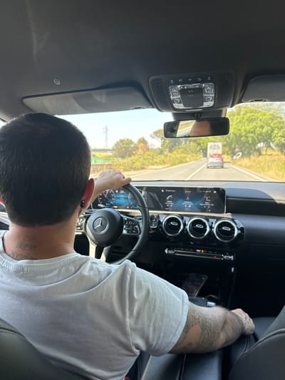 The image depicts the interior of a car, specifically the driver's seat area, with a person driving. The person has short, dark hair, wearing a white shirt and tattoos on his left arm. His hands are on the steering wheel, and the dashboard features a digital display with navigation information. The car is traveling on a road, and scenery is visible through the windshield including trees, a road, a truck and buildings. The scene suggests that the car is likely located in a suburban or rural area. Based on information from the question, this is in Ardea, Italy. The interior of the car shows a modern design with climate control vents and other dashboard features. The rearview mirror is visible in the upper center of the image.