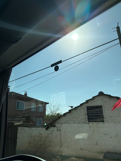 The image captures an outdoor scene, likely viewed from inside a vehicle, under bright, clear daylight conditions. The time of day appears to be mid-day due to the intense sunlight causing a prominent sun flare and rainbow-like reflections on the windshield.

**Subjects:** The primary subjects include two buildings, utility lines, a clear blue sky, and elements of a vehicle's interior. One building on the right is old, with an off-white, cracked facade and a small, recessed window opening fitted with vertical slats. A red flag, likely the flag of Turkey, is visible near its roofline. Behind and to the left, a second, darker (brown/reddish-brown) building stands, featuring multiple windows, some reflecting the sky. Utility lines or power cables stretch across the sky, with a circular marker or device attached to one. Sparse tree branches are visible behind the buildings. The foreground reveals parts of the vehicle's interior, such as a dark dashboard or roof lining and a structural frame, viewed through the windshield, which is marked by numerous reflections.

**Activity or event:** No specific activity or event is taking place; the image is a static snapshot of the environment, likely taken from a parked or moving vehicle.

**Notable details:** The intense sunlight creates significant lens flare and reflections on the glass, which distort some parts of the view. The weathered appearance and visible cracks on the white building suggest its age. The presence of a red flag strongly indicates the location is Turkey, aligning with the provided context of Iznik.

**Visible text:** A black rectangular sticker is affixed to the inside of the vehicle's windshield. It features a circular symbol resembling a speaker or bell with a diagonal line through it, and faint text below it, which appears to be "HGS" (a common automated toll payment system in Turkey).