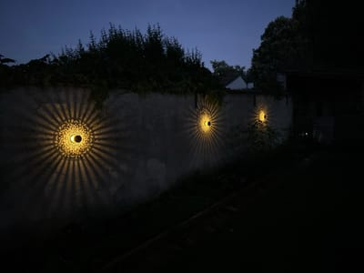 The image depicts three solar-powered wall lights mounted on a textured, light-colored stone wall. The lights are circular with intricate patterns that cast radial light and shadow designs onto the wall. They are spaced along the wall, with the leftmost light being the largest and brightest. The wall is lined with dense, dark green foliage along its top edge. Beyond the wall, glimpses of buildings, including a white-pitched roof, and mature trees are visible against a twilight sky. The ground in front of the wall is dark and appears to be a garden or yard, with a raised wooden structure or path visible in the lower right portion of the frame. The scene is set in the city of Villa Pasquali, Italy, suggesting an outdoor residential or garden setting.
