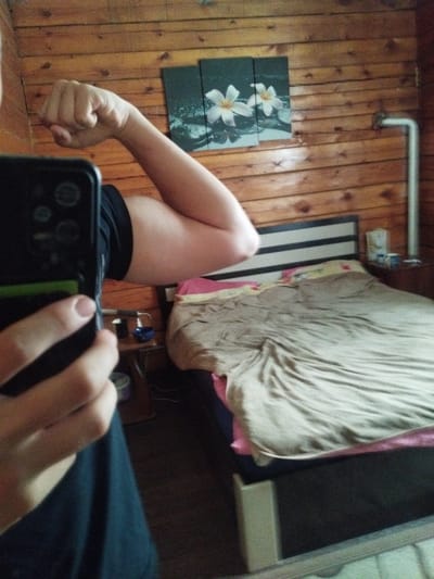 A first-person perspective shot shows an individual holding a smartphone in their left hand, angled to capture a reflection. The individual is flexing their right arm, displaying their bicep. The background consists of a wooden paneled wall. On the wall, there is a framed triptych artwork featuring two white flowers. To the right of the artwork, a white pipe runs vertically. In the reflection, a bed with light brown bedding and a pink accent is visible. A bedside table with a blue object and other items is also present. The overall scene suggests an indoor environment, likely a bedroom. The image does not provide any visual cues to determine the city of Kirensk, Russia.