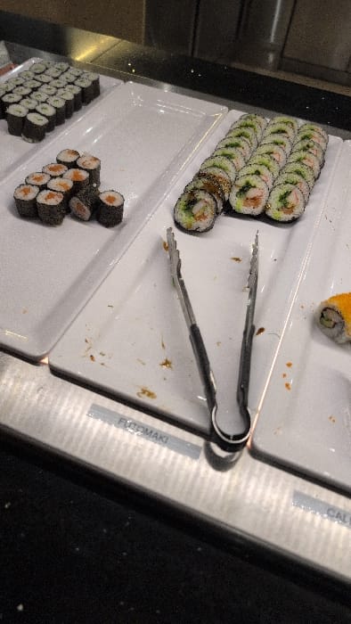 This image captures a sushi buffet display within a commercial establishment in Horst, Netherlands. The scene is dominated by three white rectangular serving dishes arranged on a stainless steel countertop.
The leftmost dish contains approximately 35-40 uniformly arranged maki rolls, each featuring a light green filling, likely cucumber, encased in dark nori seaweed. Adjacent to this, a second white dish holds approximately 10-12 maki rolls with an orange-red filling, possibly salmon or tuna. Some of these rolls are slightly dislodged, suggesting recent patron activity.
The central dish prominently displays a long, dense row of approximately 25 uramaki (inside-out) rolls, characterized by white rice exteriors and visible fillings including shrimp and green vegetables. A silver-toned pair of serving tongs with black handles rests open on this central dish. Below the dish, a grey label with white text identifies the sushi type as "FUTOMAKI."
Evidence of active use is present in the form of brownish liquid splatters and small food particles scattered across the white serving surfaces, particularly around the serving tongs. A single maki roll, adorned with bright orange roe, is partially visible on the far right edge of the frame. The foreground features a dark, textured countertop, and the background consists of dark, reflective panels. No individuals are visible within the frame.
