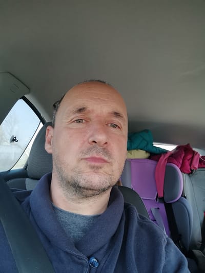 A man is in the driver's seat of a car. He is wearing a blue jacket and a grey shirt. He has a receding hairline and a stubbly beard. A seatbelt is fastened across his chest. The back seat is filled with items, including a purple child car seat, a green puffy jacket, and a pink jacket. The environment appears to be Slaveevo, Bulgaria. The location is derived from metadata embedded in the image.
