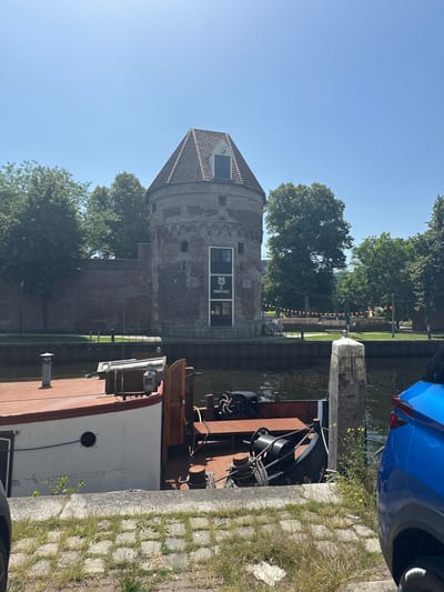 The image shows a view from a boat docked on a canal in Zwolle, Netherlands. A historic brick tower with a tiled roof, likely a part of the city's old fortifications, stands prominently on the left bank. The tower has several small windows and a larger window with text above a boarded-up section. Some green trees are visible behind the tower, and beyond them, a park area with grass, some small trees, and orange banners. The canal water is dark and reflects the surroundings. On the right side of the image, the edge of a blue car is visible, parked on a paved area next to the canal. A portion of the boat the image is taken from is also visible, showing metalwork and ropes. The scene is bathed in bright sunlight, suggesting it is daytime on a clear day. There are no people clearly visible in the image. The overall atmosphere is peaceful and historical. The text visible on the tower appears to be "DE WIJNBROEDERS".