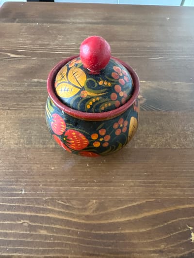 An overhead shot shows a wooden pot with a lid on a wooden table. The pot is decorated with a black background and colorful floral patterns in red, yellow, and orange. A red, spherical knob sits on top of the lid. The table is made of dark wood planks with visible grain. The background is blurred, showing hints of a lighter colored wall or surface. The scene is static and appears to be indoors. The image is in focus and well-lit. This scene is not in Balqaş, Kazakhstan.