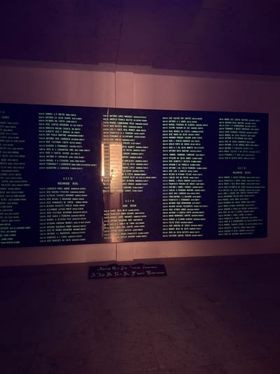 This image depicts a large, dark rectangular memorial plaque mounted on a light-colored wall, likely within a building in Praia do Ribatejo, Portugal. The plaque is filled with multiple columns of light-colored text, which lists names, each typically prefixed with "SOLD-PQ" (likely an abbreviation for "Soldado-Paraquedista," meaning Paratrooper-Soldier) followed by a name and a date (e.g., 01.09.73). Headers such as "RCP 21," "RCP 31," and "RCP 12" are visible, alongside geographical references including "MOÇAMBIQUE" (Mozambique), "GUINÉ" (Guinea), and "ANGOLA," indicating a memorial for those who served in the Portuguese Colonial War. A prominent vertical light reflection obscures parts of the text in the central columns of the main plaque. Below the large plaque, resting on a light-colored tiled floor, is a smaller, dark rectangular plaque with light-colored text, inscribed with the Portuguese phrase: "Aquilo que por terras fizemos e que da na la a Norte libertando." The scene is dimly lit with a purple/magenta hue in the shadows. No individuals are present.