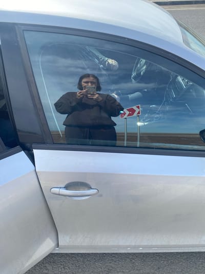 Here's a detailed description of the image:
**Scene:** The image shows the door of a silver-colored car. A person, presumably the photographer, is reflected in the car window, holding a smartphone and taking a picture. The background visible through the window suggests an outdoor environment.
**People:** One person is visible as a reflection. Their appearance is partially obscured by the reflection, but they appear to be holding a smartphone.
**Objects:** Key objects include the silver car, specifically its door, the car window, a smartphone held by the reflected person, a road sign.
**Environment:** The road sign, reflecting the light, appears to be in Kliuchiki, Russia. The sign indicates a direction and is along a roadside.