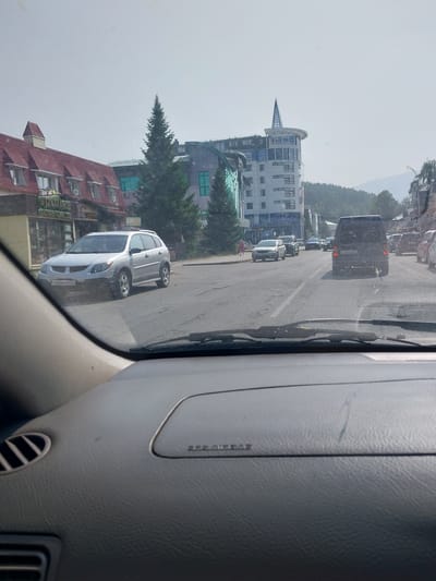The image depicts a street scene in Belokurikha, Russia, viewed from the interior of a car. In the foreground, the dashboard and windshield of the car are visible. The street shows moderate traffic, including a silver Pontiac Vibe in the left lane and a dark-colored van in the right lane. Buildings line the street, with a prominent multi-story building with a distinctive pointed roof in the background. One building on the left has a red pitched roof and signage that appears to be in Russian. The environment appears to be a developed area with trees and mountainous terrain visible in the distance under a hazy sky. A pedestrian is seen walking on the sidewalk. The time of day appears to be daytime, with bright lighting conditions.