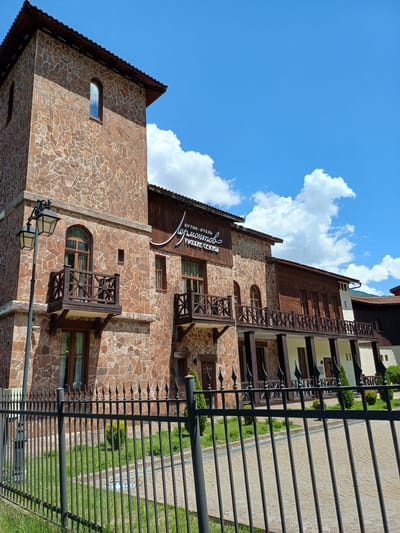 The image displays the exterior of a multi-story building complex, identified as "БУТИК-ОТЕЛЬ Лермонтов РУССКИЕ СЕЗОНЫ" (Boutique Hotel Lermontov Russian Seasons), located in Эстосадок, Russia. The building features a prominent tower-like section on the left, constructed from irregular brown and reddish-brown stone masonry, with dark-framed arched windows. The main structure extends to the right, incorporating additional stone masonry sections, dark wooden paneling on upper floors, and multiple dark wooden balconies with decorative railings. A continuous balcony supported by dark pillars spans a portion of the lower floor. The roof is dark and sloped. In the foreground, a black metal fence with pointed finials runs across the scene, separating a strip of green grass from a paved walkway made of light-colored pavers. A tall, black, double-headed street lamp stands between the fence and the building on the left. Small green shrubs are visible in the landscaping. The scene is illuminated by bright sunlight under a clear blue sky with scattered white cumulus clouds. Distant green hills are visible on the horizon to the right. No individuals are present in the image.