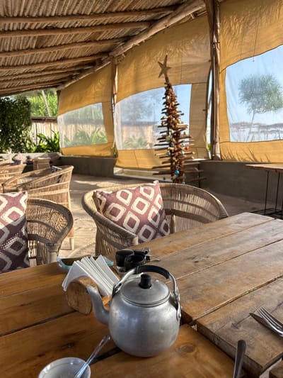 This image captures a rustic and inviting outdoor or semi-outdoor dining area, likely a cafe or restaurant in Kiwengwa, Tanzania, illuminated by bright daylight. The scene is characterized by natural materials and a relaxed ambiance.
In the foreground, a robust wooden table occupies the right side of the frame, set with a polished silver kettle, a stack of white napkins in a wooden holder, and cutlery including forks and spoons. A small camera and a white cup with a gold rim are also visible on the table.
Behind the table, several chairs made of woven natural fibers are arranged, each adorned with a cushioned pillow featuring a geometric pattern in purple and off-white. The seating suggests a comfortable and laid-back environment.
The overhead structure consists of a canopy system combining opaque, light-colored fabric sections with translucent panels, allowing natural light to filter in and offering glimpses of the sky and distant green trees. Wooden beams and a wall constructed from vertical wooden or reed slats further enhance the natural, tropical aesthetic.
A unique decorative piece hangs from the canopy: a vertical structure crafted from small wooden sticks, shells, and colorful beads, resembling a bohemian-style Christmas tree or a large mobile, topped with a star ornament.
The image conveys a sense of peaceful relaxation, typical of a holiday destination. No people are present, emphasizing the calm and serene atmosphere. There is no visible text in the scene.