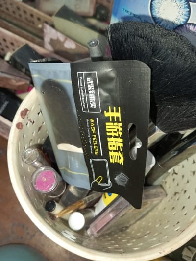 A white, round, woven basket holds various cosmetics and makeup tools. Prominently featured is a black package labeled "WASP FEELERS Mobile Gaming Finger Sleeve," with Chinese characters above the English text. The package also displays an image of a hand using a smartphone. Other items in the basket include lipsticks, brushes, and miscellaneous cosmetic containers. A blue contact lens case can be seen in the background.