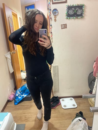 A young woman is captured taking a mirror selfie in an indoor setting, likely a bedroom or hallway. She is wearing a long-sleeved black top, black jeans, and white socks. Her long, dark brown hair cascades over her shoulders, partially obscuring her face as she holds a smartphone with a patterned case to capture her reflection. A ring is visible on her right-hand ring finger.
The room features a light-colored wall decorated with various items, including a framed picture of two white swans on a dark background with a green and white fringe border, a smaller framed image, a decorative plate, and a carved wooden spoon. Below the mirror, a wooden shelf is visible, upon which a small metal fixture and some plastic bags rest.
The wooden floor shows a white digital bathroom scale, several pairs of sneakers (including black, green, and black with pink details), and multiple plastic bags. A prominent blue shopping bag has the text "YOYÓ" visible on its side. The overall scene suggests a personal and lived-in space, illuminated by what appears to be natural daytime light.