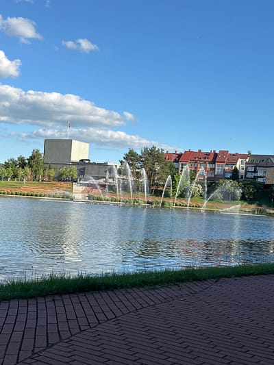 The image depicts an outdoor scene featuring a canal with multiple active water fountains. A paved walkway made of dark red bricks runs along the right side of the canal, bordered by a strip of green grass. On the left side of the canal, there is a park-like area with trees, manicured bushes, and a modern building with a light-colored facade and a red flag. Further in the background, several residential buildings with red-tiled roofs are visible. The sky is a clear blue with scattered white clouds. The sun appears to be shining, casting reflections on the water's surface, suggesting it is daytime. The overall atmosphere is serene and well-maintained, indicating a public space. There is no visible text in the image. The location context provided is Panevėžys, Lithuania.