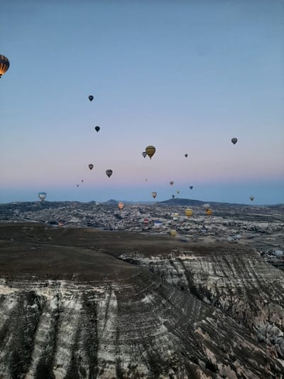 An aerial shot captures a landscape dotted with numerous hot air balloons, likely in Cappadocia, Turkey, near the city of Avanos. The scene unfolds under a dusky sky, with the horizon transitioning from a pale blue to a soft pink hue. In the foreground, a rugged, layered terrain with distinct white and dark brown striations dominates the lower portion of the frame, suggesting a geological formation. Scattered across the middle ground and extending into the background, a collection of stone structures and buildings form a settlement. The sky is populated by a multitude of hot air balloons, varying in size, color, and pattern, some illuminated, drifting in different directions and altitudes. The perspective suggests the photo was taken from another hot air balloon.