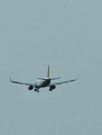 The scene captures a twin-engine commercial passenger aircraft in mid-flight over Norderstedt, Germany. The aircraft is viewed from its rear and slightly beneath its path, centered against a uniform, light blue-gray sky. Its landing gear is deployed, indicating a state of approach or controlled descent. The fuselage is white, complemented by a yellow or orange vertical stabilizer. Two engines are visible beneath the wings, which are equipped with winglets. The environment consists solely of the unfeatured sky; no other subjects or objects are present within the frame.