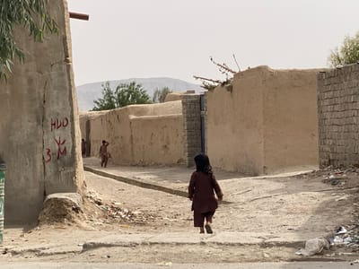 A young girl wearing a brown tunic walks down a narrow dirt street lined with mud brick walls. The street leads to a small alley.  Two other children are visible in the distance, walking down the alley toward a small tree.  The street is covered in debris and litter. The location is a residential area in Panjwai, Afghanistan.  A large, cracked mud brick wall with graffiti and a metal pipe sticking out is visible in the foreground.  The building on the opposite side of the street has a brick wall and a crumbling mud brick wall. The sky is clear and blue with no clouds.