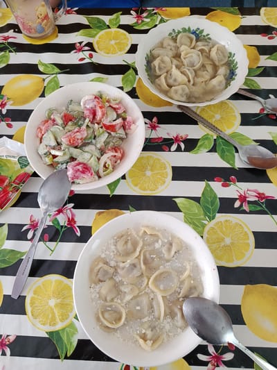 A close-up, overhead shot shows a table set with two bowls of pelmeni (dumplings) and a bowl of salad. The table is covered with a black and white striped tablecloth adorned with illustrations of lemons and pink flowers. To the left, a glass mug with a picture of Winnie the Pooh holding a pot of honey sits on a coaster. One bowl of pelmeni is in the upper right quadrant, and the other is in the lower center. The salad, made with chopped tomatoes, cucumbers, and onions dressed in a creamy sauce, is in a bowl in the upper left quadrant. Several spoons are placed around the bowls. The scene is indoors, likely a dining area. The image does not provide any specific information about the location in the city of Koylyk, Kazakhstan.