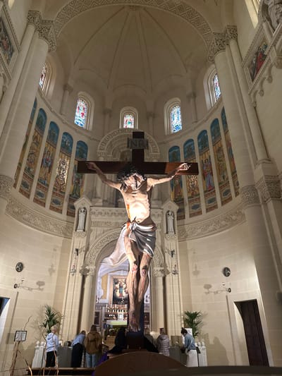 The image depicts the interior of a large church in Għarb, Malta. A prominent, life-sized statue of Jesus Christ on a dark wooden crucifix is centrally positioned in the foreground, facing the viewer. The statue shows Christ with a pale complexion, a white loincloth, and visible red wounds on his hands, feet, and torso. The inscription "INRI" is affixed above his head on the cross.

The church interior is constructed of light-colored stone, featuring a high, vaulted ceiling and a central dome with intricate carved patterns. Numerous tall, arched stained-glass windows, displaying colorful religious figures and designs, are set into the upper walls. Below these windows, a series of arched panels contain mosaic artworks depicting angels, flora, and religious symbols. A continuous band of ornate relief carving runs along the lower walls, which also contain niches holding white statues of saints.

In the background, an ornate altar is visible, featuring a painting of the Virgin Mary and child, flanked by numerous lit candles and decorative elements. To the left of the crucifix, a smaller shrine or altar is present, adorned with a green potted plant. A wooden stand nearby holds a sign reading "TAIS FEGĦRET / SILENZIO / SILENCE / STILL."

Several individuals are present within the church. On the left, three people stand near the smaller shrine; one person in a light blue hooded sweatshirt faces the shrine, while two others in dark and brown jackets stand nearby. On the right, one person in a dark top is seated on a pew, and another individual in a light-colored jacket and skirt stands facing the front of the church. The lighting is a combination of natural light from the stained-glass windows and artificial illumination, casting shadows on the textured walls.
