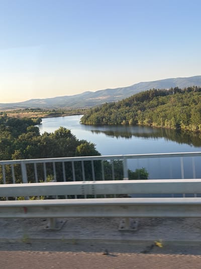 A photograph captures a tranquil landscape featuring a winding river, dense green forests, and distant rolling hills under a clear blue sky. In the foreground, a metallic guardrail runs horizontally across the image, suggesting the vantage point is from a bridge or elevated roadway. The river, a calm expanse of dark water, reflects the surrounding trees and the sky. The vegetation is lush, with various shades of green indicating a mix of trees and foliage. The background hills are softer in color, blending into the hazy atmosphere. There are no people or visible signs of urban development in the scene. The overall impression is one of natural beauty and serenity.