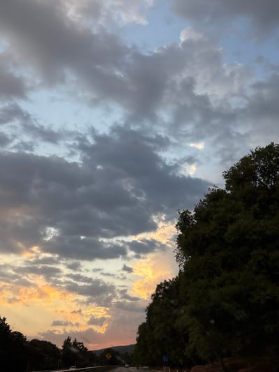 The image depicts a view of the sky with clouds, some illuminated by the setting sun, creating an orange hue. A line of trees is visible along the right edge, suggesting a landscape or roadside setting. In the bottom right corner, a glimpse of a road with a road sign can be seen. The overall scene appears to be a natural environment with some man-made elements like the road. The location is Tepoztlán, Mexico.