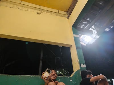 The image depicts two young individuals at night, likely outdoors, given the glimpse of a tiled roof and exposed wooden beams above. One person, in the foreground and on the left, is a child with dark hair who is smiling broadly while holding a smartphone. Their body is partially visible, suggesting they are sitting or leaning. The setting appears to be a slightly dilapidated building with yellow and green painted walls. The yellow wall has a rough, unfinished texture along the top edge, revealing brickwork underneath. The green wall has white specks, possibly paint splatters or imperfections.

The second person, on the right side of the frame, is a young male with dark hair, wearing a black t-shirt. He is leaning against the wall, with his arms around himself, and appears to be looking downwards or away from the camera. The overall mood is casual and relaxed, with the smiling child in the foreground adding a touch of joy. The time of day is clearly night, indicated by the darkness outside the window and the artificial lighting, which casts reflections and creates lens flares in the upper right section of the image. The location context of Barengkrajan, Indonesia, suggests a tropical or warm climate where outdoor evening activities are common. No visible text is present in the image.