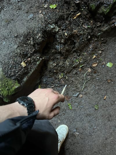 A person is holding a lit cigarette in their right hand, appearing to be in an outdoor, possibly wooded, area. The ground consists of dirt, leaves, and small branches. A section of the ground is dark and has some moss. The person is wearing a black jacket and a black digital watch. A white shoe is visible in the background. It is dark, suggesting nighttime. There is no information to suggest the location is Garraunbaun, Ireland.