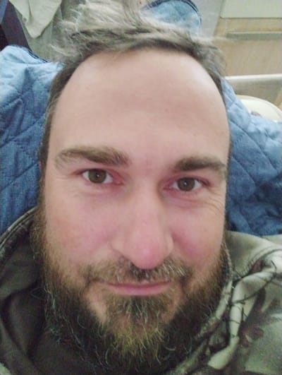 The image is a close-up, selfie-style photograph of a Caucasian man with a beard and light brown hair. He is looking directly at the camera with a neutral expression. He is wearing a camouflage jacket. The background appears to be indoors, with a quilted blue fabric behind his head and upper body, possibly a blanket or bedding. The lighting is soft and appears to be artificial. There is no discernible indication of the man's location in Konstantinovo, Russia, or any other specific location.