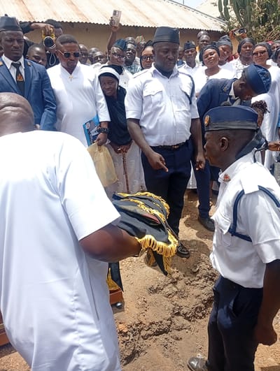 A solemn outdoor gathering is depicted, featuring a large crowd of people under bright daylight in what appears to be an unpaved area, consistent with Ladura Foron, Nigeria. Many attendees are men dressed in various uniforms; some wear dark blue suits with black hats and ties, while others sport white short-sleeved shirts with dark blue trousers and similar hats. Several women are also present, some wearing black head coverings.
In the foreground, a man with his back to the viewer, dressed in a white traditional garment, holds a black and gold ceremonial item, possibly a folded uniform or regalia, featuring elaborate gold fringing. To his right, a man in a white short-sleeved uniform shirt with a black hat observes the scene.
Central to the image is a man wearing a white long-sleeved garment, sunglasses, and a prominent gold cross necklace. He holds a blue brochure or document that clearly displays "70" and "2023" along with an image of a person in uniform, suggesting an event program or memorial booklet. Near him, an older woman with a black head covering appears somber, holding a walking stick. In the background, simple structures with corrugated iron roofs are visible. The overall atmosphere, attire, and the ceremonial item strongly suggest a formal event, likely a funeral or memorial service. Expressions on the faces of the crowd are predominantly serious or solemn.