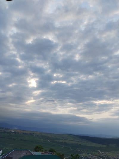 The image depicts a cloudy sky over a landscape featuring rolling green hills and a few scattered buildings. A bright patch of sunlight breaks through the clouds in the upper center of the frame. The lower portion of the image shows rooftops and glimpses of what appear to be rural or semi-rural structures, including what might be a greenhouse or shed with a green roof. The hills in the middle ground are covered in vegetation, possibly vineyards or fields, and stretch towards distant, hazy mountains. The overall impression is of an overcast day in a somewhat remote, scenic area.