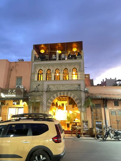 The image depicts a street scene in Mechouar Kasbah, Morocco, featuring the Kasbah Cafe. The cafe is a multi-story building with traditional Moroccan architectural details, including arched windows and intricate patterns. The cafe is illuminated with warm light emanating from within, creating a welcoming atmosphere. A yellow car is parked in the foreground, partially obstructing the view of the cafe's entrance. A scooter is parked in front of the building, suggesting a lively street scene. The sky is transitioning from day to night, casting a blue hue over the scene. A sign for a business named "KOK" is visible below the Kasbah Cafe.
